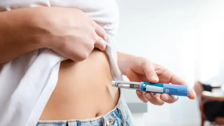 Person lifting shirt to self-administer a subcutaneous injection with a blue pen device into the abdomen