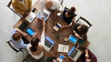 Overhead shot of a business meeting at a wooden table with open laptops and a handshake at the center